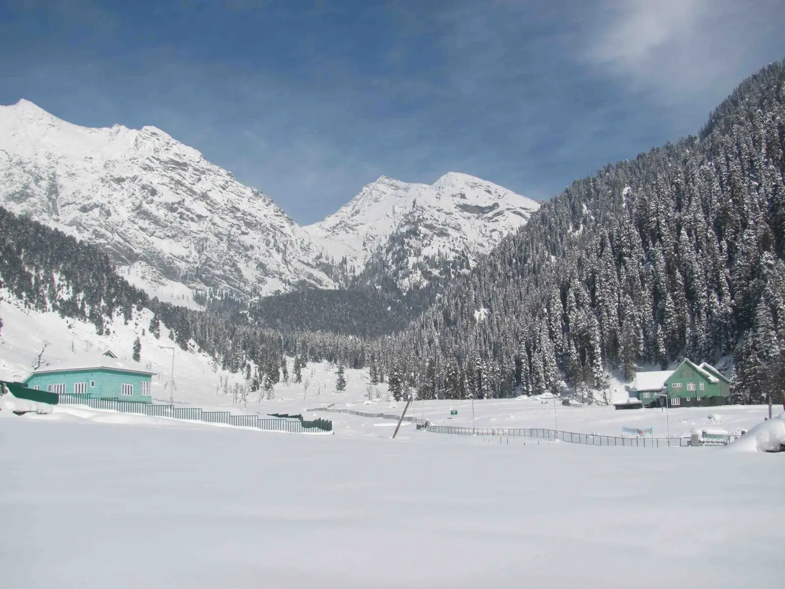 Aru Valley near Pahalgam under deep winter snow, with snow-laden pine forest and two green-roofed huts below white peaks