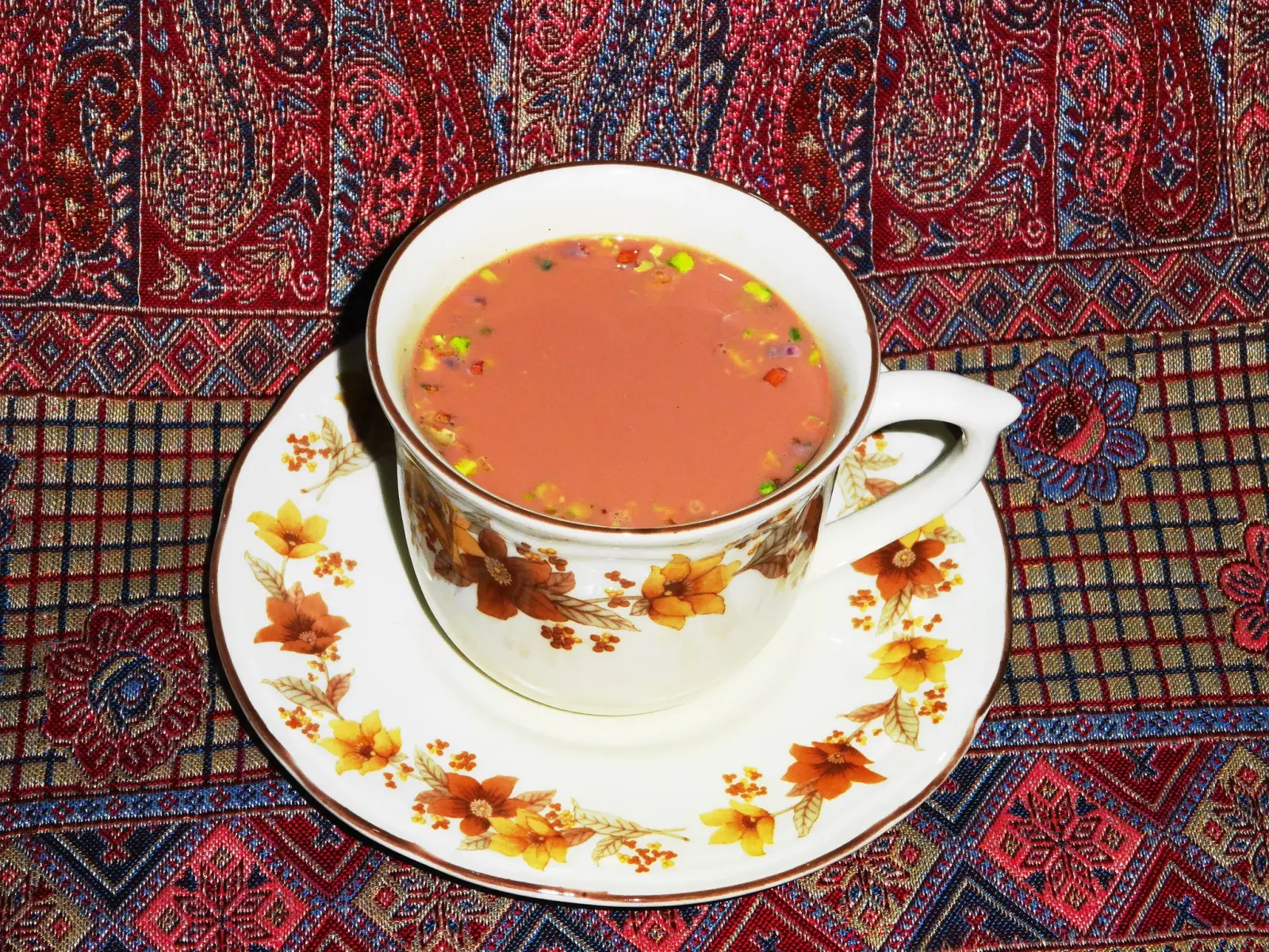 A cup of pink Kashmiri tea with crushed nuts on a floral saucer, resting on a red woven Kashmiri textile