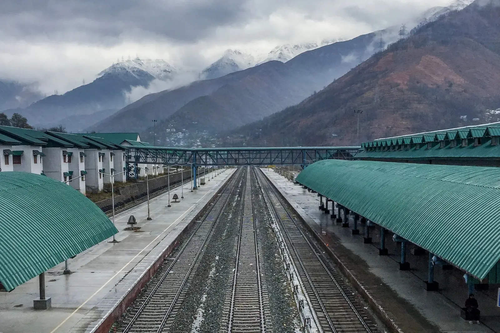 Banihal railway station in Jammu and Kashmir, with empty platforms under green canopies and snow-capped mountains beyond