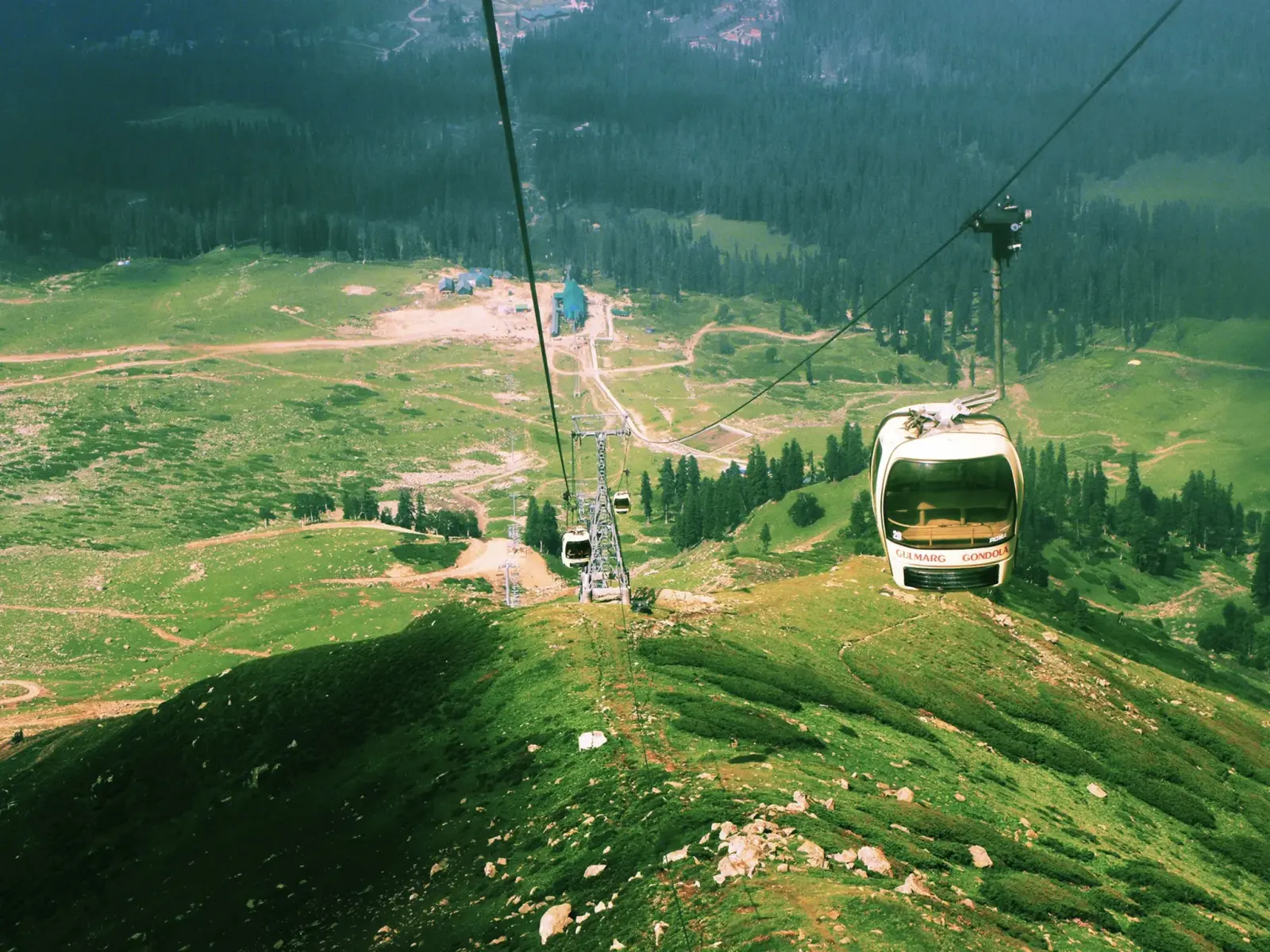 Gulmarg gondola cabins on their cable above a green summer meadow and pine forest in Gulmarg