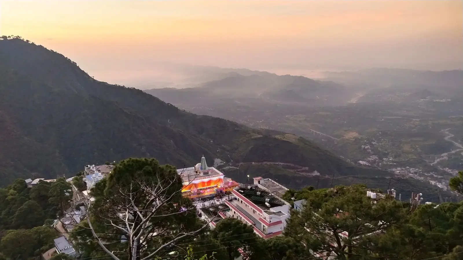 Katra town and the Trikuta foothills at twilight, with lit buildings among trees and hazy hills fading into the distance