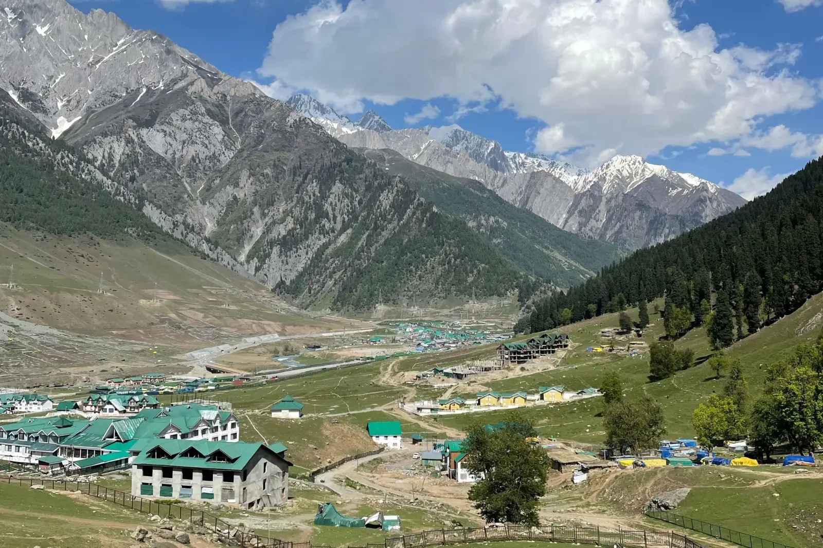 Sonamarg valley in summer with green slopes, a cluster of buildings and rugged snow-streaked mountains under blue sky
