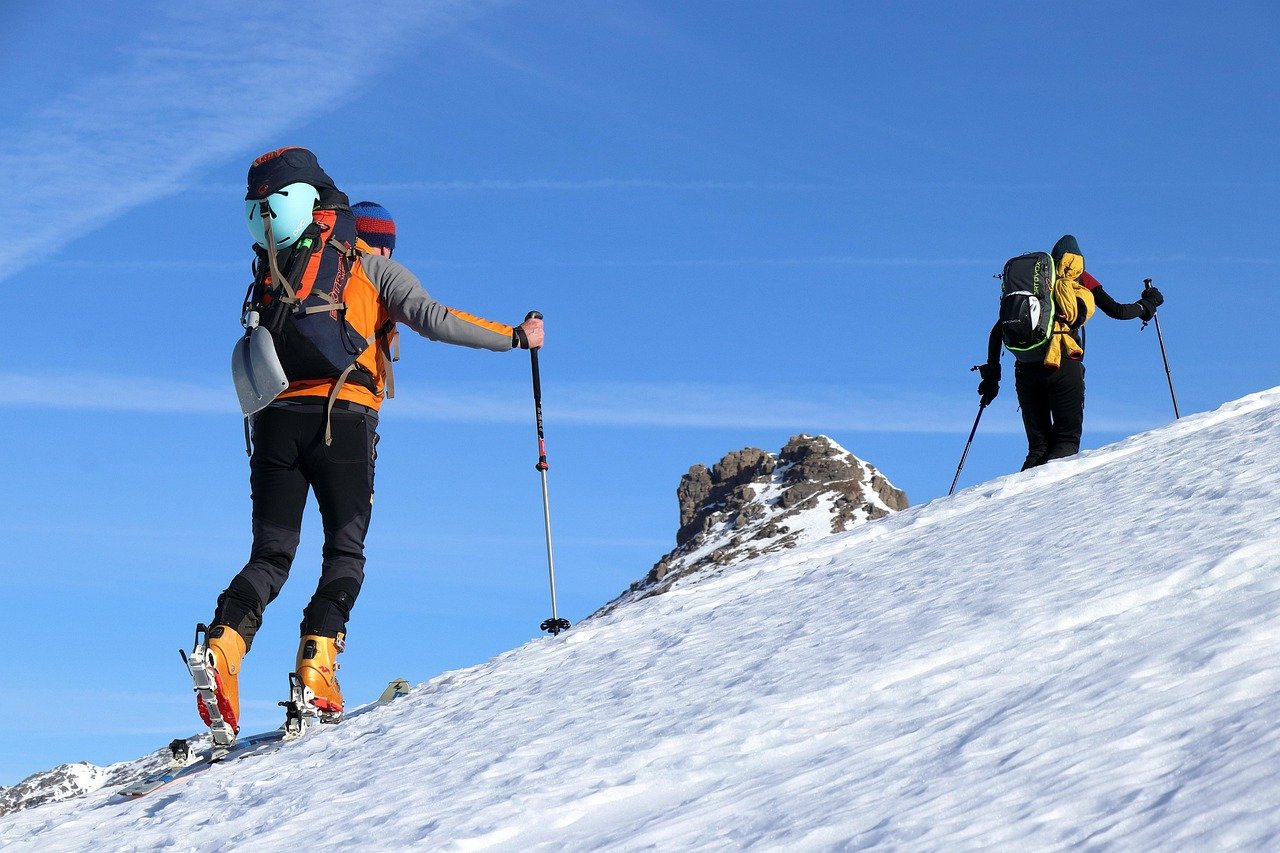 Two backcountry skiers with touring gear climbing a steep snow slope under a clear blue sky