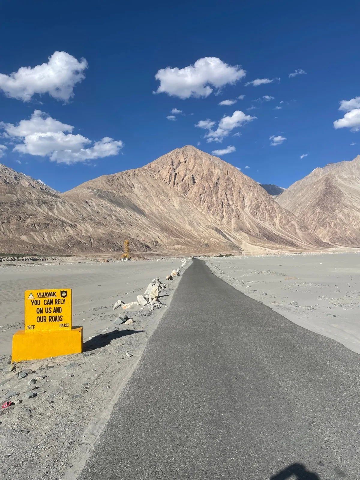 A paved mountain road cutting through the high-altitude desert terrain of Ladakh, bare brown peaks under a clear blue sky with scattered clouds, the kind of landscape the Srinagar-Leh highway crosses into once past Kargil