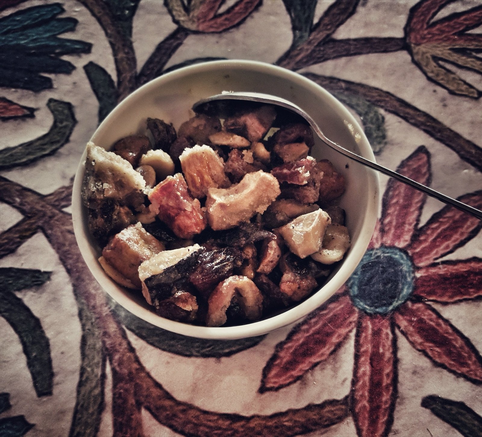 A copper bowl of Kashmiri shufta on a traditional dastarkhwan (floor tablecloth) -- the warm amber-gold sweet visible in the foreground, the mixed dry fruits and golden paneer cubes distinguishable in the dish, a light scatter of dried rose petals across the surface, a small silver bowl of saffron-coloured syrup beside it, in the background the dastarkhwan extends with other Kashmiri dishes, the brass serving utensils resting on the cloth edge, late afternoon interior light