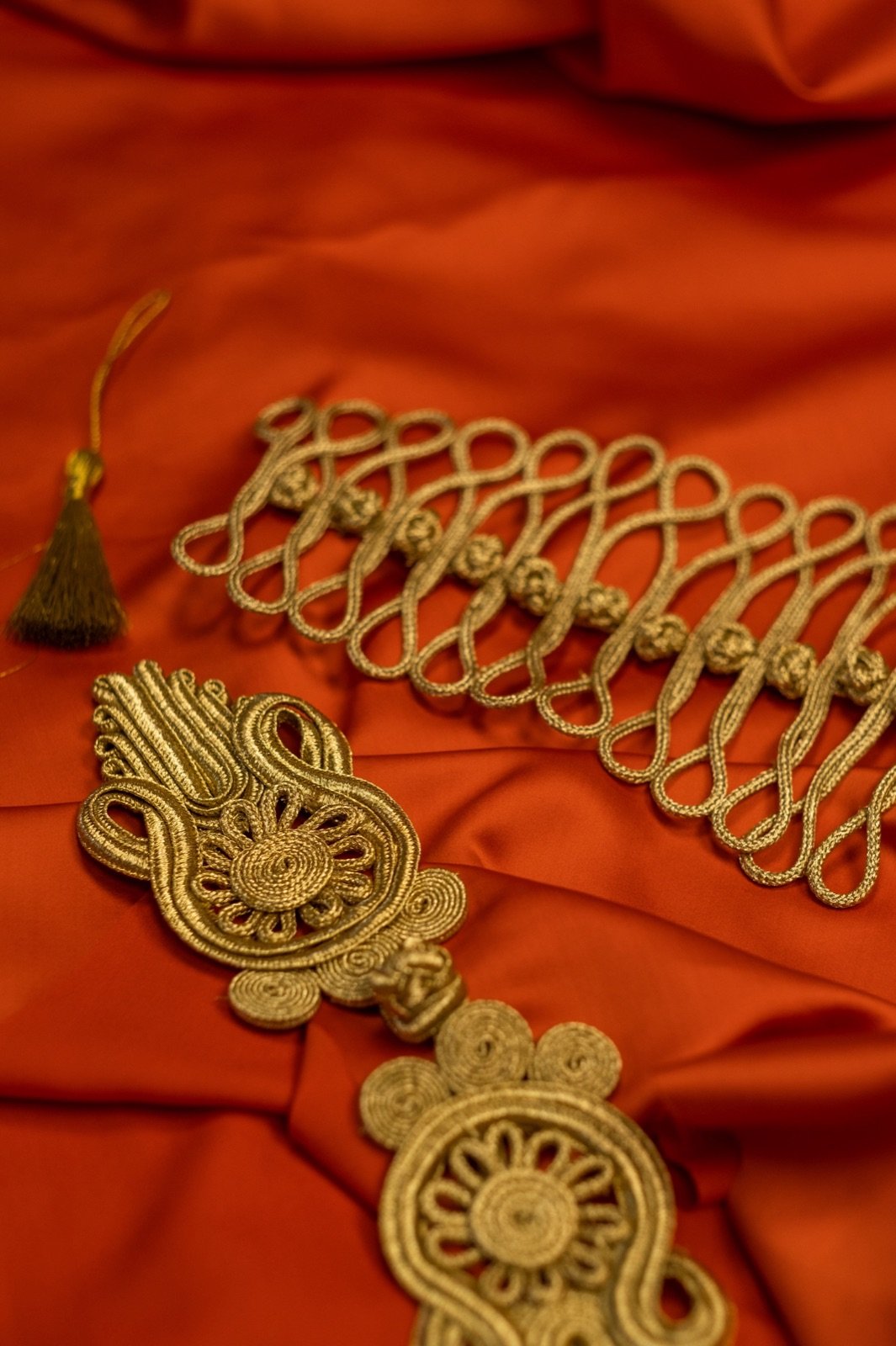 A craftsperson working Tilla Daraz in a Srinagar Old City workshop -- seated on the floor with a large frame holding dark red velvet, the ari hook visible in their right hand, a reel of metallic gold thread in the foreground, narrow window light illuminating the partially completed floral pattern