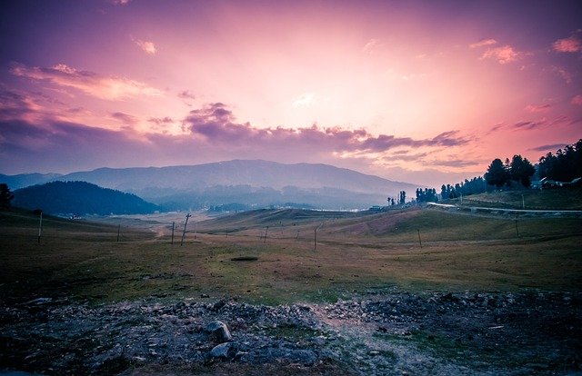 Gulmarg meadow in late September -- the open grassland showing the first transition from deep green to amber and gold, the Apharwat ridge behind with a light dusting of early snow on the upper rocky face, a handful of visitors visible at distance on the meadow, the Gulmarg Gondola cable visible against the pale September sky, morning light at a low angle across the meadow, no crowds, the fir treeline at the meadow edge beginning to show the first hint of autumn colour
