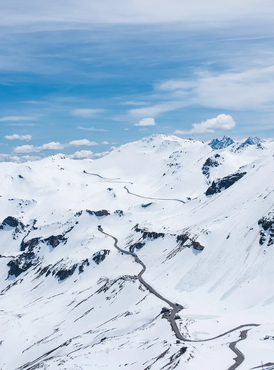 Snow-covered mountain highway road in winter, deep fresh snow on both sides of the road, high mountain peaks with heavy snowfall visible in the background, dramatic Himalayan winter pass scenery