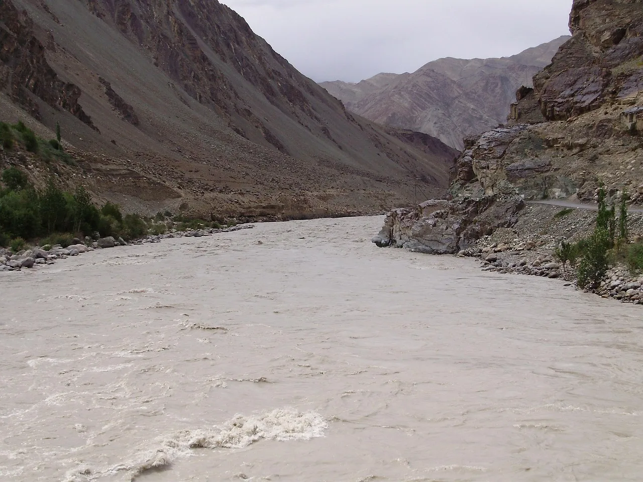The road approaching Chandanwari in June -- the Lidder River running fast in the narrow gorge below, pine-covered slopes rising steeply on both sides, a white snowfield visible at the upper end of the valley where the road terminates, a few vehicles parked at the snowfield edge
